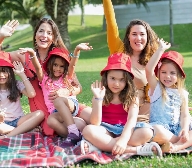 Two women and four girls enjoy a sunny picnic in Portugal, waving happily in the park.